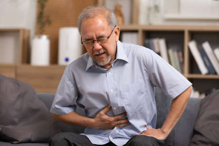 Senior men holding his stomach feeling pain while sitting on the sofa at home. Asian man having stomachache with isolated gastric. Senior suffering from digestion problem or acid reflux.の写真素材