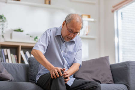 Asian old man with his knee joint pain in sofa, pain in the elderly, health care, elderly care. Elderly man having a knee pain and sitting down. Grandfather with knee pain.の写真素材