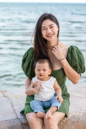 Happy cute baby boy with his mother on the beach. Summertime with baby is a quality time for Asian mommy.  Mother and baby on the beach and happy morning moment. Motherhood love and care for her childの写真素材