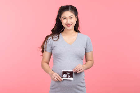Happy Pregnant Woman standing smile holding ultrasound image stroking big belly with love isolated on pink background,Pregnancy of young woman enjoy with future life,Motherhood and Pregnant Conceptの写真素材