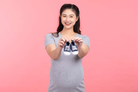 Asian pregnant woman holding baby sneakers for baby newborn isolated on pink background. Pregnant...