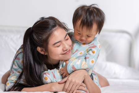 Cute Asian mom hugging her baby boy on bed smile and happiness together. Little baby toddler sit on bed and relax with mother speding time to learning and growth. Good momen of family time. Mother dayの写真素材