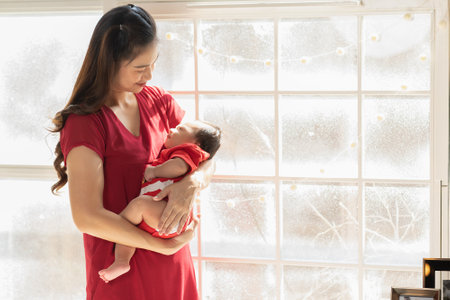 Beautiful Asian mom woman wear red cloth holding newborn baby in arms standing looking outside...