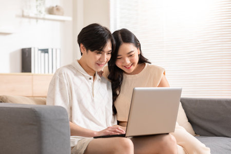Asian young couple sit on couch or sofa looking at Computer Laptop smile and laughing together. Cheerful lover surfing internet and social media spending time in holiday at cozy home. Valentine Dayの写真素材