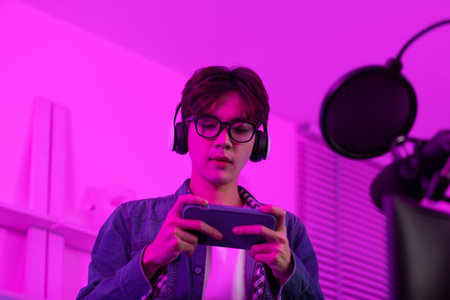 Professional Asian gamer playing and live streaming online game by smartphone in game room. Young man streaming video game on mobile phone with headphones and smiling, enjoying online gaming and tech.の写真素材