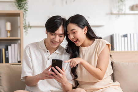 Asian young couple sit on couch or sofa looking at mobile phone smile and laughing together. Cheerful lover surfing internet and social media spending time in holiday at cozy home. Valentine Dayの写真素材