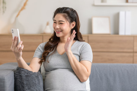 Happy Pregnant Woman sitting on  couch video call conference on mobile phone with family at cozy home.Pregnancy of young woman enjoying with future life relax at homeの写真素材