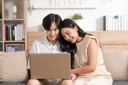 Asian young couple sit on couch or sofa looking at Computer Laptop smile and laughing together. Cheerful lover surfing internet and social media spending time in holiday at cozy home. Valentine Dayの写真素材
