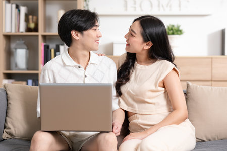 Asian young couple sit on couch or sofa looking at Computer Laptop smile and laughing together. Cheerful lover surfing internet and social media spending time in holiday at cozy home. Valentine Dayの写真素材