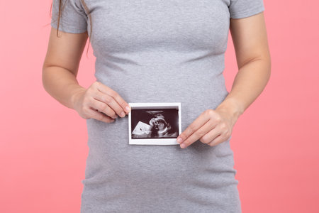 Happy Pregnant Woman standing smile holding ultrasound image stroking big belly with love isolated on pink background,Pregnancy of young woman enjoy with future life,Motherhood and Pregnant Conceptの写真素材