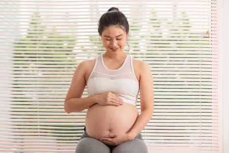 Happy Pregnant Asian Woman sitting on a bed holding and stroking her big belly at home. Pregnancy of young woman enjoying with future life relax at home. Motherhood and Pregnant Concept. Soft focusの写真素材