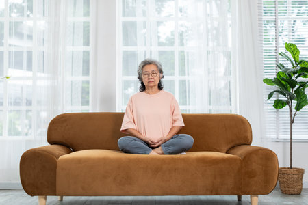 Mature Asian woman doing yoga for meditation on sofa in living room at home.Clam of senior or elderly women meditation deep breath and relax at cozy home.Happy Retired Meditation at home conceptの写真素材