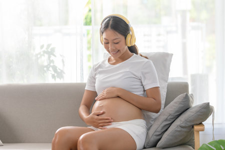 Pregnant woman sitting on couch, listening to music with headphones, hand on belly, wearing comfortable clothes, relaxed and happy at home.の写真素材