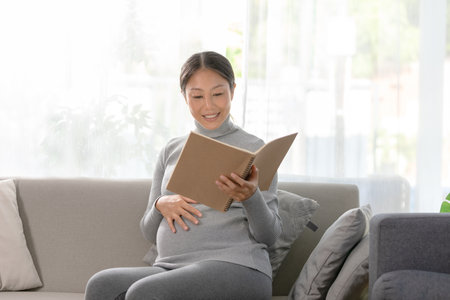 Asian pregnant woman is seated on a comfortable sofa, engrossed in reading a book. She gently touches her belly, Serene and reflective moments of pregnancyの写真素材