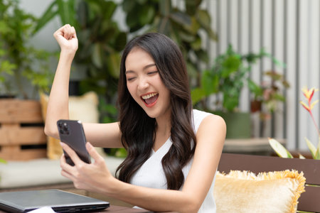 Millennial asian young woman looking mobile phone laughing with good news or discount voucher for shopping online at home.Happy and cheerful woman looking on cellphone app read message feel excitedの写真素材