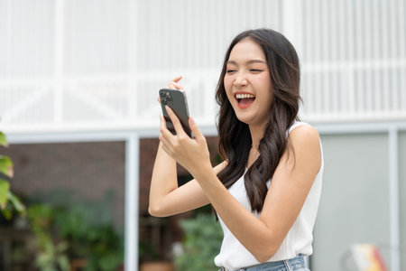 Millennial asian young woman looking mobile phone laughing with good news or discount voucher for shopping online at home.Happy and cheerful woman looking on cellphone app read message feel excitedの写真素材
