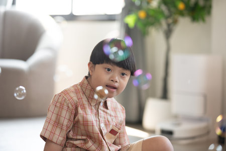Asian cheerful young boy with Down syndrome enjoys playing with bubbles indoors. His bright smile...