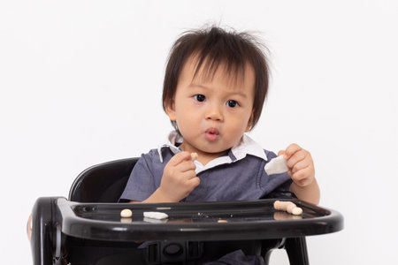 Asian adorable baby sits in a high chair, holding snacks in both hands. The baby wears a blue outfit with a white collar and has an inquisitive expressionの写真素材