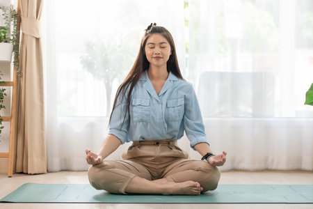 Asian young woman meditating in a calm home environment. Seated cross-legged on a yoga mat, she exudes tranquility and focus, representing themes of mindfulness, wellness, and inner peaceの写真素材