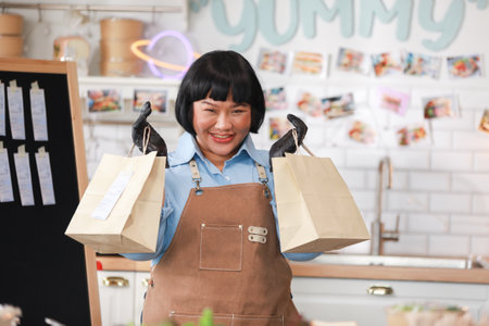 Cheerful Asian chef in brown apron and black gloves proudly holds up two takeaway bags, ready for delivery. The kitchen background is vibrant and cozy. Small business conceptの写真素材