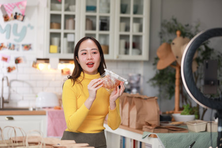 Female vlogger in bright yellow sweater enthusiastically presents food product in cozy kitchen. Captured mid-presentation through ring light, with smartphone positioned in front of her for recordingの写真素材