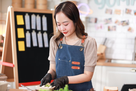 Cheerful young female chef, dressed in blue apron and black gloves carefully prepares sandwich in cozy kitchen. Kitchen warmly decorate with various cooking utensils and ingredients visible on counterの写真素材