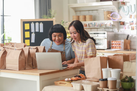 Two cheerful Asian chefs share a joyful moment as they look at a laptop in a cozy kitchen. Surrounded by takeaway bags and delicious food items, they exhibit a sense of accomplishment and happinessの写真素材