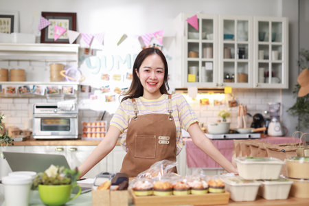 confident woman entrepreneur stands proudly in her kitchen, surrounded by prepared food items neatly packaged for sale. She wearing apron and smiling, exuding sense of accomplishment and professionalの写真素材