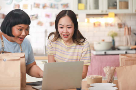 Two cheerful Asian chefs share a joyful moment as they look at a laptop in a cozy kitchen. Surrounded by takeaway bags and delicious food items, they exhibit a sense of accomplishment and happinessの写真素材