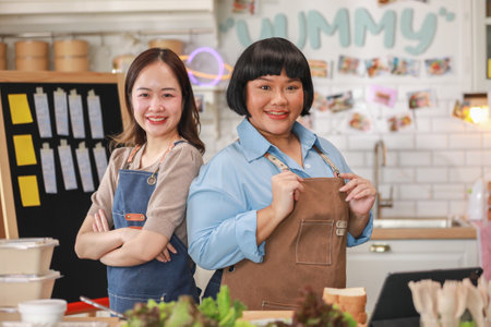 Asian two confident female chefs stand side by side in a lively kitchen, both smiling warmly. Dressed in aprons, they exude professionalism and teamwork. Small Business Conceptの写真素材
