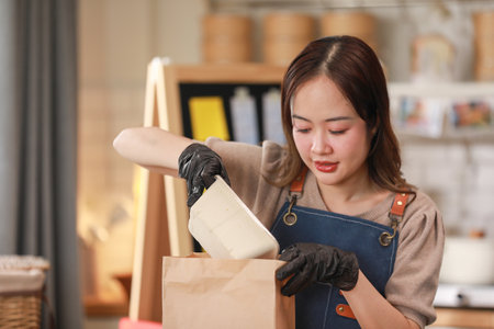 Asian young female chef, wearing apron and gloves, carefully packs food into paper bag in kitchen. Chef focused expression highlights her attention detail and care in preparing the meal for takeawayの写真素材