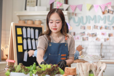 Asian young female chef, wearing an apron and gloves, carefully prepares a fresh salad in a lively kitchen environment. The background features colorful decorations and a display board. Small businessの写真素材