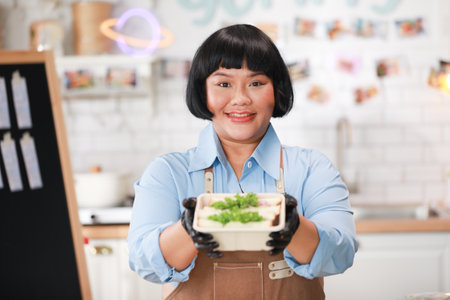 Cheerful Asian chef wearing a brown apron and black gloves presents a freshly prepared sandwich box, smiling warmly at the camera. Small Business Conceptの写真素材