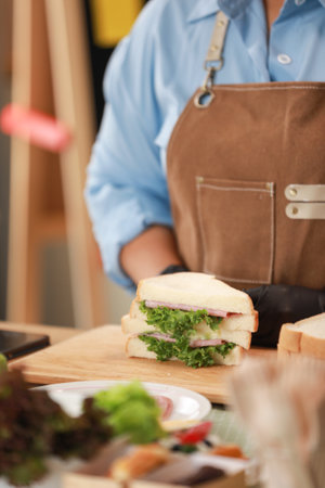 close-up view of a freshly prepared sandwich placed on a wooden cutting board. The sandwich, filled with lettuce, ham, and cheese, is cut in half, showcasing the layers of fresh ingredientsの写真素材