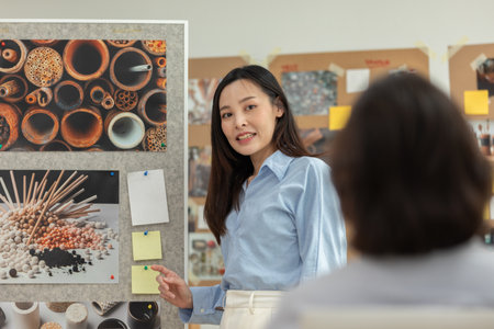 Asian professional woman delivers presentation to her colleagues during creative meeting. She gestures toward bulletin board filled with visuals and notes, explaining key ideas and strategiesの写真素材