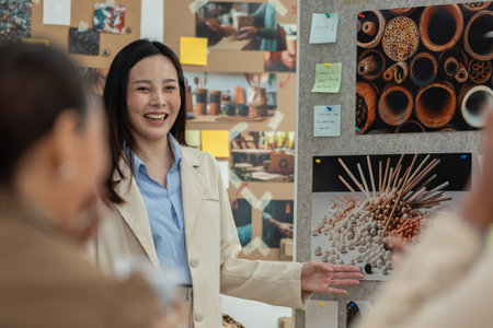 Asian professional woman smiles while leading creative presentation during team brainstorming session. She gestures toward bulletin board filled with visuals and notesの写真素材