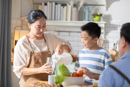 Heartwarming scene of Asian grandmother and her grandson in the kitchen. The grandmother is pouring milk into a glass while the grandson watches with smile. Happiness momentの写真素材