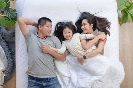 Asian joyful family scene with father, mother, and daughter lying together on bed. They are smiling and looking at each other, sharing moment of closeness and warmthの写真素材