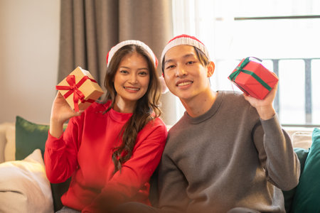Cheerful Asian young couple lover both wearing red and white holiday hats, smiles while holding wrapped gifts. Woman dressed in red sweater and man in gray,They seated indoors on cozy couchの写真素材