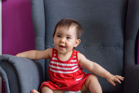 Little baby sitting on home bedroom armchair with short-sleeved red dressの写真素材