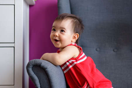Little baby sitting on home bedroom armchair with short-sleeved red dress