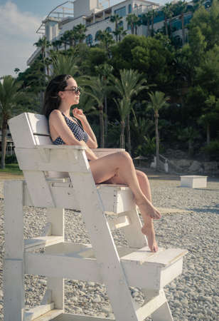 Girl sitting in a lifeguard chair wearing sunglasses touching her hair while sunbathingの写真素材
