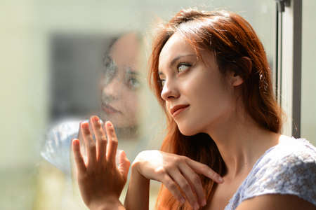 redhead girl looking through the window, worried woman. quarantined woman, woman looking through...