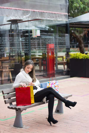 Vertical shot of a satisfied Hispanic woman sitting on a bench looking into shopping bagsの写真素材