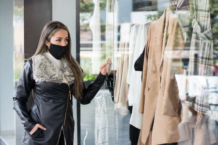 A stylish elegant Hispanic woman with a face mask looking at a shop display through the windowの写真素材