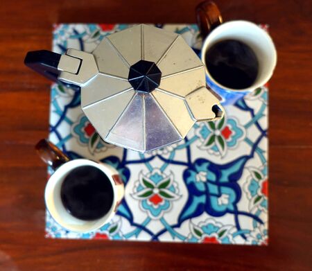 A ready-made mocha coffee machine with two full cups resting on a colorful tile on a wooden tableの写真素材