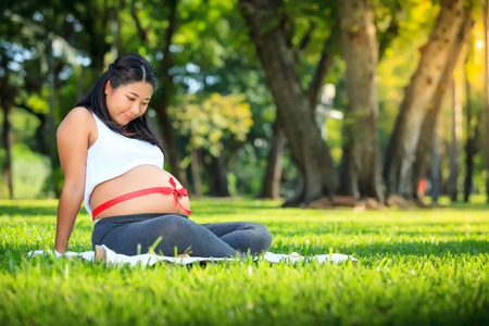 Beautiful pregnant asian woman relaxing in the park with red ribbonの写真素材