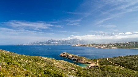 Blue skies over the The citadel of Calvi with the maquis of La Revellata in the foreground, a deserted beach and snow capped mountains in the distanceの写真素材