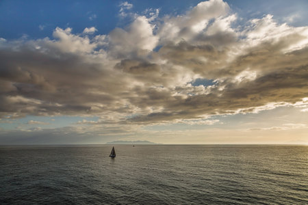 Two sailing boats pass a silhouetted island of Capri with blue skies and clouds aboveの写真素材