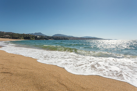 Waves gently lapping onto the beach at Propriano with golden sand, blue sea and blue sky and hills in the backgroundの写真素材